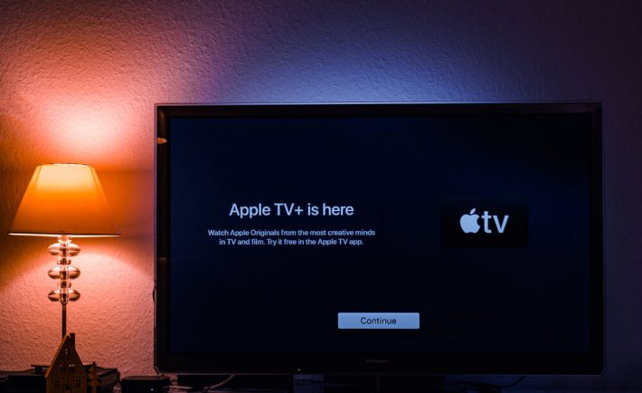widok telewizora z włączonym Apple TV, na tle białej ściany z kolorowym oświetleniem