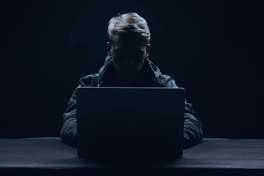postać hakera siedzącego za laptopem. Ciemne kolory.