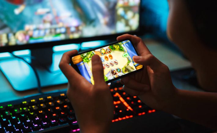Spojrzenie zza pleców na telefon, na którym ktoś gra w gry. W tle komputer, również z włączoną grą oraz podświetlana klawiatura gamingowa.