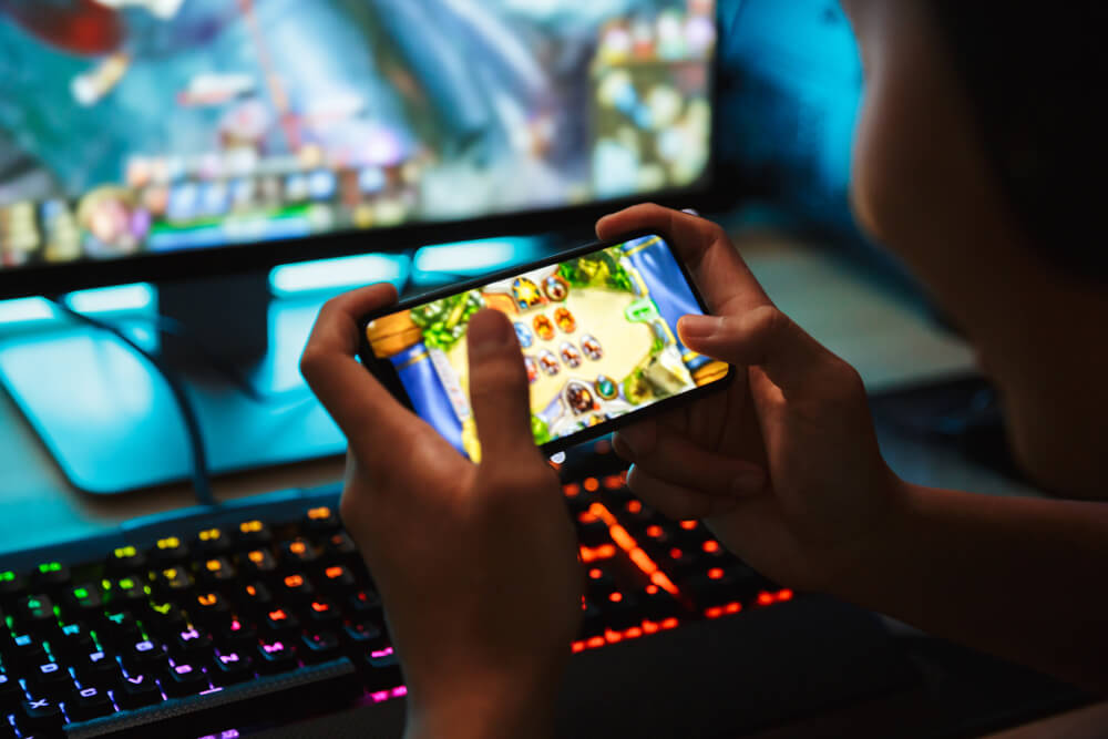 Spojrzenie zza pleców na telefon, na którym ktoś gra w gry. W tle komputer, również z włączoną grą oraz podświetlana klawiatura gamingowa.