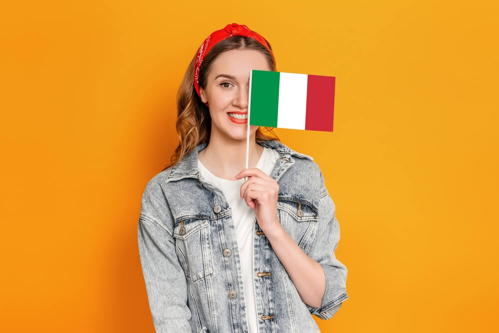 Dziewczyna zasłaniająca sobie jedno oko flagą włoską. Stoi na pomarańczowym tle.