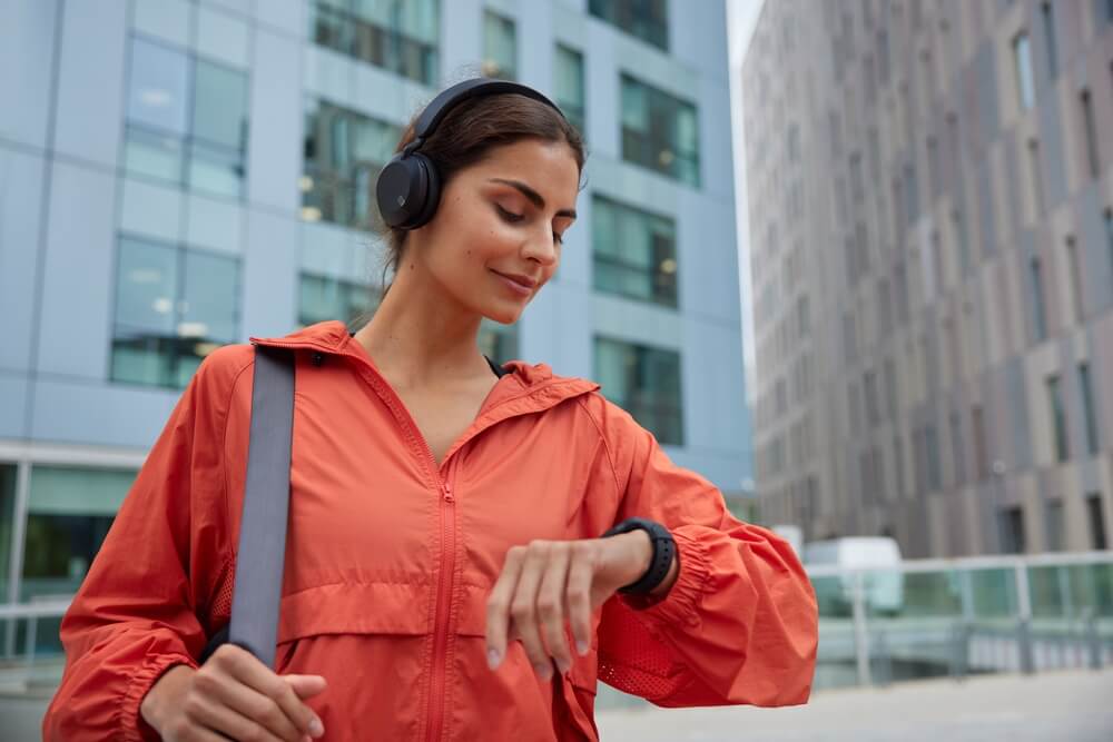 Kobieta ubrana w pomarańczową bluzę ortalionową, ze smartwatchem na ręku i w słuchawkach.